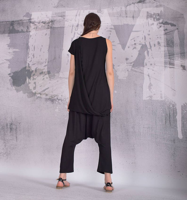 Puede incluir: Un top negro de un solo hombro con un dise&ntilde;o drapeado y pantalones de har&eacute;n negros a juego. La modelo lleva el atuendo sobre un fondo gris con salpicaduras de pintura blanca.