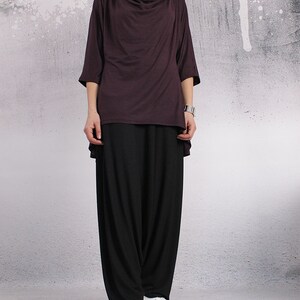 Puede incluir: Una mujer lleva una blusa larga y holgada de color morado oscuro con cuello de pico y pantalones negros anchos. Lleva zapatillas blancas.