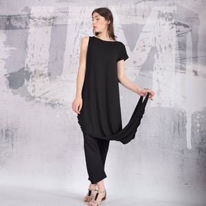Puede incluir: Un vestido negro de manga larga con un dobladillo asim&eacute;trico drapeado. El vestido est&aacute; hecho de una tela suave y fluida y tiene un ajuste holgado. La modelo lleva el vestido con pantalones negros y sandalias.