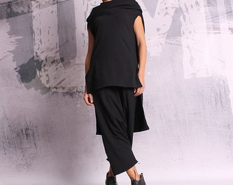 May include: A woman is wearing a black outfit consisting of a sleeveless top with a draped neckline, wide-leg pants, and ankle boots. The top has an asymmetrical design. The boots have laces and a wedge heel. The background is a textured gray and white wall.