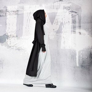 May include: A woman wearing a long, black hooded coat over a gray maxi dress. She is walking towards the camera with a white wall in the background.