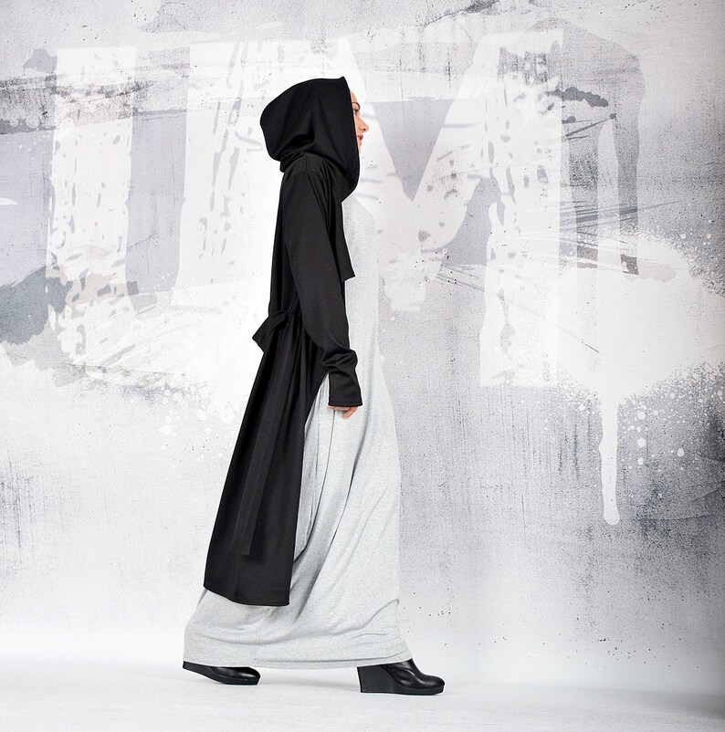 May include: A woman wearing a long, black hooded coat over a gray maxi dress. She is walking towards the camera with a white wall in the background.