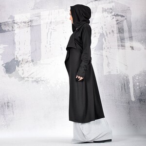 May include: A long black hooded coat with a draped front and long sleeves. The coat is worn over grey pants.