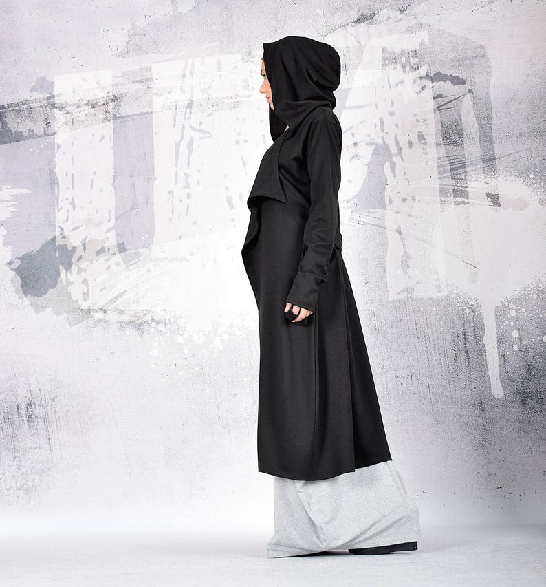 May include: A long black hooded coat with a draped front and long sleeves. The coat is worn over grey pants.