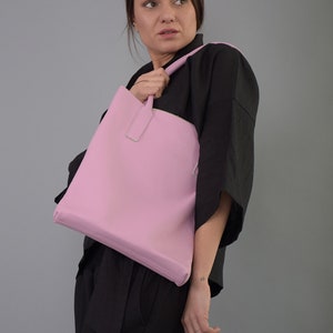 May include: A pink leather tote bag with a top handle. The bag is being held by a person wearing a black top.