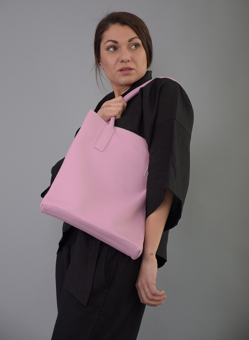 May include: A pink leather tote bag with a top handle. The bag is being held by a person wearing a black top.