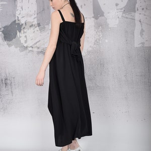 May include: A black maxi dress with wide straps and a bow detail at the back. The dress is paired with white and gray patterned platform shoes. The model is standing in front of a gray and white textured wall.
