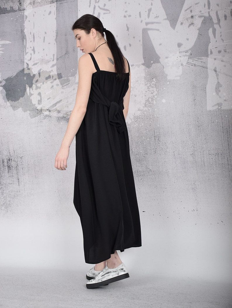May include: A black maxi dress with wide straps and a bow detail at the back. The dress is paired with white and gray patterned platform shoes. The model is standing in front of a gray and white textured wall.