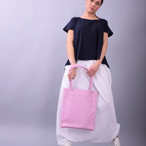 May include: A woman wearing a white skirt and a navy blue t-shirt is holding a pink leather tote bag with the initials "LM" on the front.