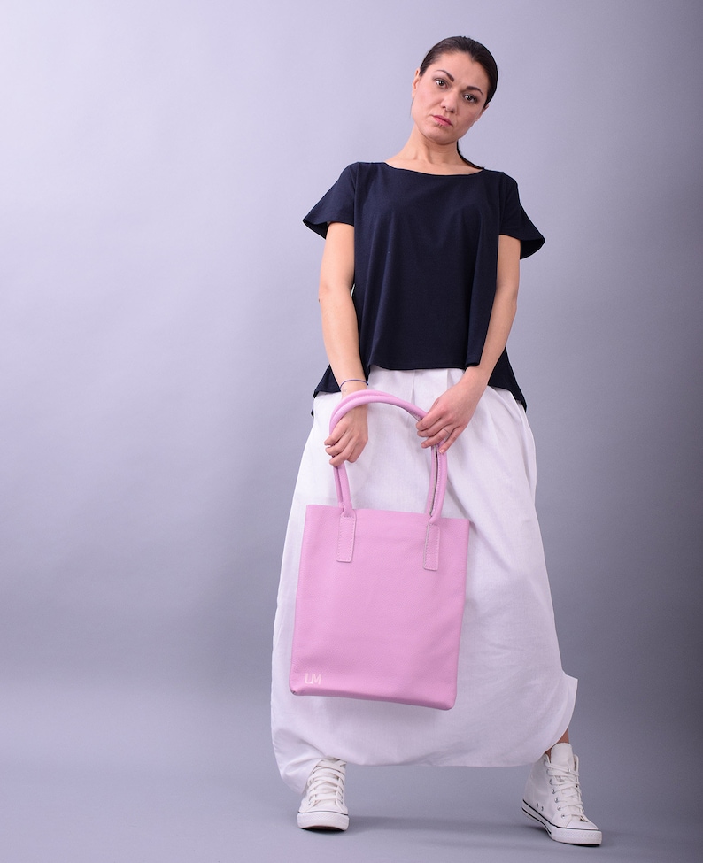May include: A woman wearing a white skirt and a navy blue t-shirt is holding a pink leather tote bag with the initials "LM" on the front.