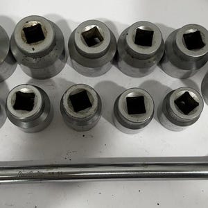 May include: A set of eleven silver-toned metal socket wrench attachments of varying sizes arranged in two rows, accompanied by a long, straight metal breaker bar handle with a knurled grip and a hinged square drive head at the end.