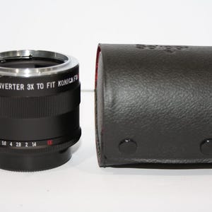 May include: A black 3x teleconverter lens with silver accents, designed to fit Konica F cameras, sits next to a black leather case. The lens has a series of numbers and markings. The case has a textured surface.
