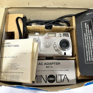 May include: A silver Minolta DiMAGE E201 digital camera with a 2.3-megapixel resolution, packaged in its original box. The box also contains an AC adapter, instruction manuals, and a black camera strap. The packaging includes text about being made of recycled paper.