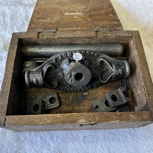 May include: A vintage Armstrong pipe threading tool set housed in a rectangular wooden box. The central cast iron die stock features the embossed text F. ARMSTRONG BRIDGEPORT CT. Several metal die inserts and a threaded pipe segment are included.