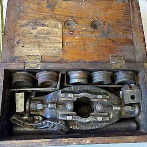 May include: A vintage wooden tool chest containing a cast iron pipe threading tool, multiple metal die heads, and a small adjustable wrench. The interior lid features a paper label with the text Please Take Notice and The Armstrong Mfg Co.