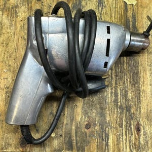 May include: A silver electric drill with a black cord wrapped around it. The drill has a chuck on the end and a trigger switch on the side.