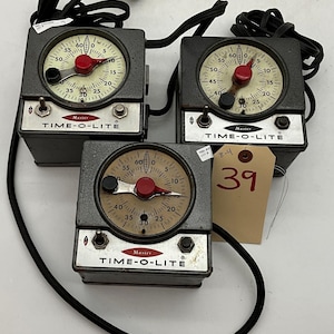 May include: Three dark gray metal Time-O-Lite Master darkroom timers with black power cords. Each unit features a circular clock face with a red center knob, a black pointer, and a silver control panel with a toggle switch and a push button.