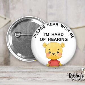 May include: A white button with the text "PLEASE BEAR WITH ME I'M HARD OF HEARING" and a cartoon bear illustration. The button has a metal pin back and is likely a pinback button or badge.