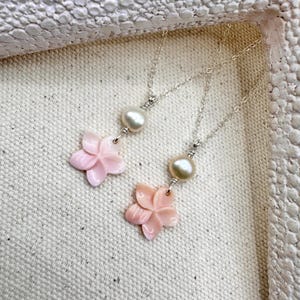 May include: Two necklaces with silver chains. Each has a light pink flower pendant and a white pearl. The necklaces are displayed on a textured, off-white surface.
