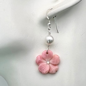 May include: A dangling earring featuring a carved light pink five-petalled flower charm with a small central accent, suspended below a single white spherical bead on a silver-toned metal hook wire, showcasing a delicate and symmetrical floral design.