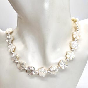May include: A necklace featuring a series of white, iridescent, carved mother-of-pearl flower beads alternating with small, round, white pearls. The floral-shaped components are arranged in a single strand, creating a uniform, light-colored design.