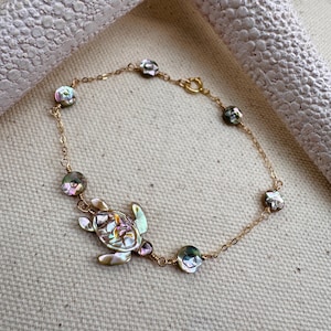May include: A gold-colored chain bracelet featuring a sea turtle charm and iridescent, round beads. The sea turtle charm has a colorful, abalone shell design. The bracelet is displayed on a neutral-colored surface.