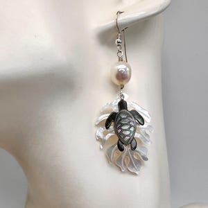 May include: Dangle earrings with a pearl and sea turtle design. The earrings have a silver hook and a large, round pearl. Below the pearl is a leaf-shaped pendant with a black and iridescent sea turtle. The leaf is white with a pearlescent sheen.