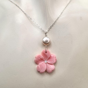 May include: A silver chain necklace featuring a pendant with a single white pearl bead and a carved light pink five-petaled flower. The center of the flower is accented with a small, clear, iridescent stone, and the entire piece hangs from a silver link chain.