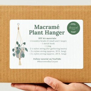 Peut inclure: Une boîte en carton marron avec une étiquette blanche qui indique "Macramé Plant Hanger" et liste les matériaux du kit DIY : 4 perles en bois (3 petites et 1 grande), 1 crochet en métal, 1 anneau, 2 x ficelle en nylon (pour les nœuds de rassemblement), 3 x ficelle en nylon (environ 3 m de long), et 3 x ficelle en nylon (environ 6 m de long). L'étiquette comprend également le texte "Suivez le tutoriel sur YouTube" et "#BurrowedbyCaryne".
