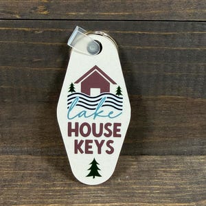 May include: A cream-colored key tag with the words "lake HOUSE KEYS" in teal and burgundy. The tag features a cabin graphic with trees and water. A clear plastic loop and metal ring are attached.