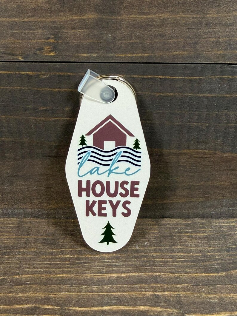 May include: A cream-colored key tag with the words "lake HOUSE KEYS" in teal and burgundy. The tag features a cabin graphic with trees and water. A clear plastic loop and metal ring are attached.