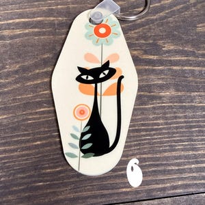 May include: A cream-colored keychain with a black cat design. The cat has a long tail and is surrounded by orange and pink flowers with green leaves. The keychain has a silver ring attached to a clear plastic loop.