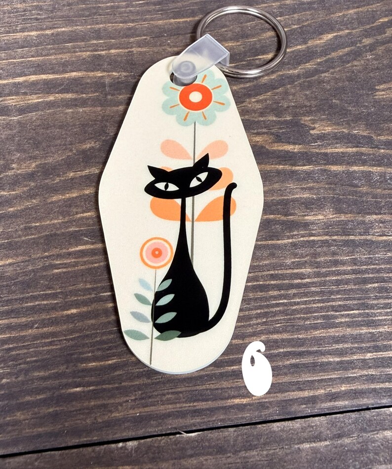 May include: A cream-colored keychain with a black cat design. The cat has a long tail and is surrounded by orange and pink flowers with green leaves. The keychain has a silver ring attached to a clear plastic loop.