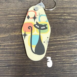 May include: A retro-style keychain featuring a black cat silhouette with a long tail, set against a cream background with colorful geometric shapes. The keychain has a metal ring and a plastic clasp for attaching keys.