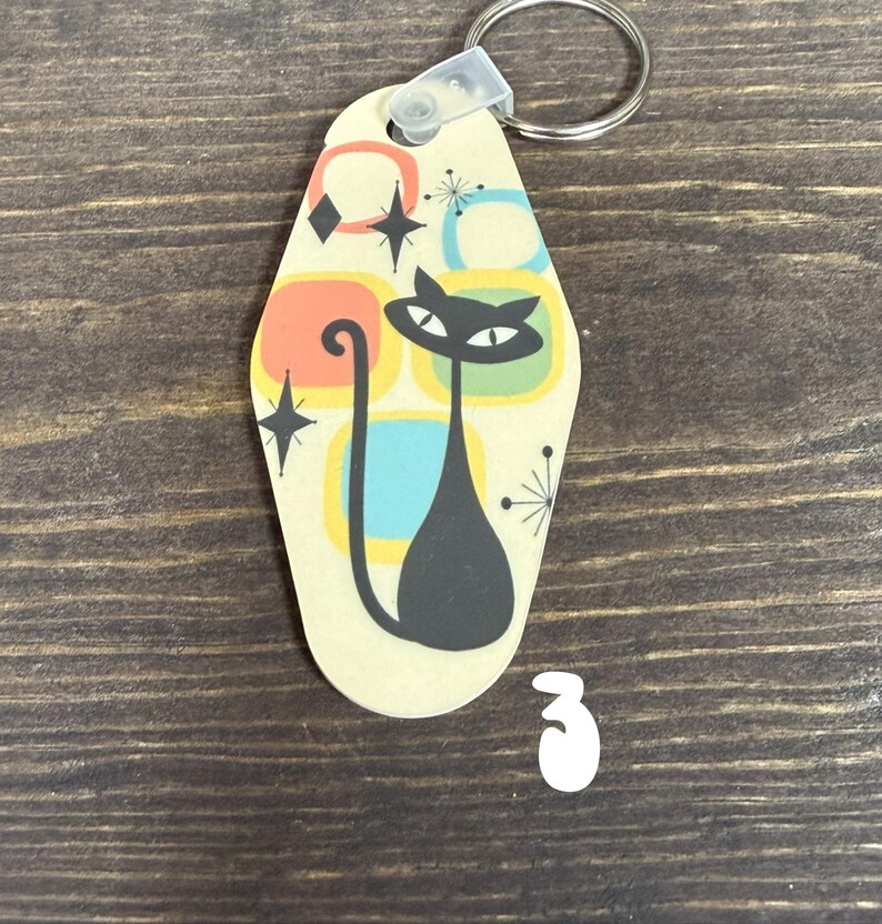 May include: A retro-style keychain featuring a black cat silhouette with a long tail, set against a cream background with colorful geometric shapes. The keychain has a metal ring and a plastic clasp for attaching keys.