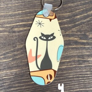 May include: A retro-style keychain with a black cat design. The keychain has a cream-colored background with mid-century modern-inspired shapes in orange, blue, and brown. A silver key ring is attached at the top.