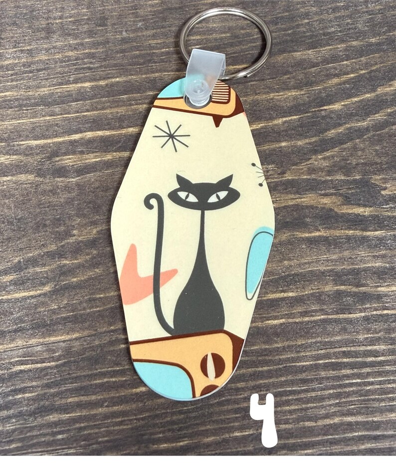 May include: A retro-style keychain with a black cat design. The keychain has a cream-colored background with mid-century modern-inspired shapes in orange, blue, and brown. A silver key ring is attached at the top.