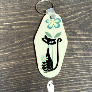 May include: A retro-style keychain with a black cat design. The keychain has a light green background with a black cat silhouette, flowers, and a metal ring for attaching keys. The number 6 is visible at the bottom.