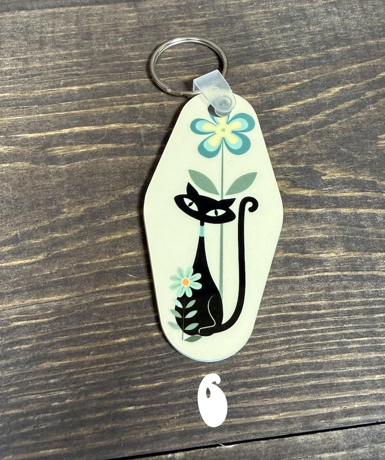 May include: A retro-style keychain with a black cat design. The keychain has a light green background with a black cat silhouette, flowers, and a metal ring for attaching keys. The number 6 is visible at the bottom.