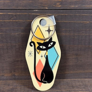 May include: A retro-style keychain featuring a black cat design. The keychain has a cream-colored background with geometric shapes in orange, yellow, and blue. A silver key ring is attached at the top. The cat has a yellow collar.