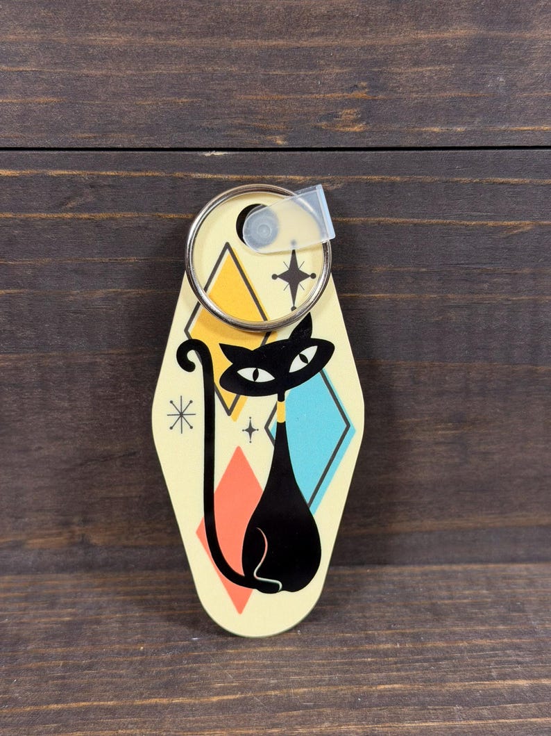 May include: A retro-style keychain featuring a black cat design. The keychain has a cream-colored background with geometric shapes in orange, yellow, and blue. A silver key ring is attached at the top. The cat has a yellow collar.