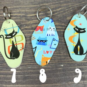 May include: Three retro-style cat keychains with silver key rings. The keychains feature black cat silhouettes and colorful geometric designs on a light green and blue background. Numbers 7, 8, and 9 are printed at the bottom.