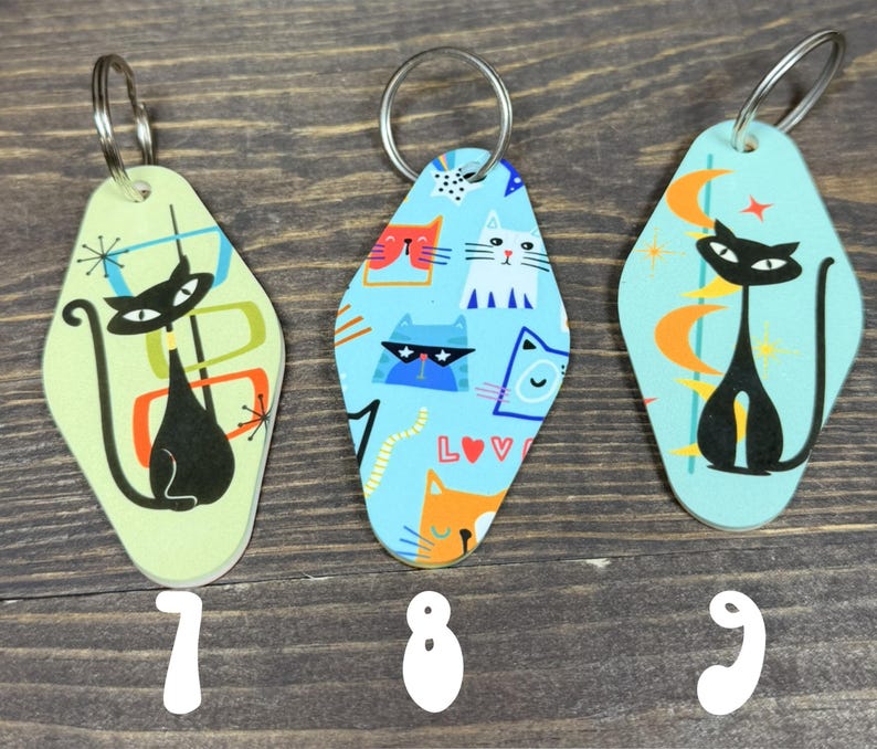 May include: Three retro-style cat keychains with silver key rings. The keychains feature black cat silhouettes and colorful geometric designs on a light green and blue background. Numbers 7, 8, and 9 are printed at the bottom.