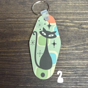 May include: A retro-style keychain featuring a black cat design. The cat has large eyes and a long tail, set against a light green background with geometric shapes and starburst accents. The keychain has a metal ring.