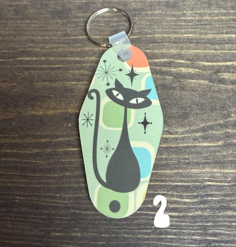 May include: A retro-style keychain featuring a black cat design. The cat has large eyes and a long tail, set against a light green background with geometric shapes and starburst accents. The keychain has a metal ring.