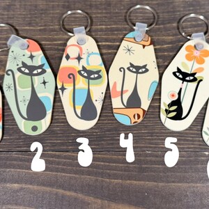 May include: Six retro-style keychains with a black cat design. Each keychain features a different background pattern with colors like orange, blue, and yellow. The keychains have a silver keyring attached.
