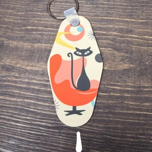 May include: A retro-style keychain featuring a black cat sitting in an orange chair. The keychain has a cream-colored background with a mid-century modern design, including a circular shape with yellow and blue accents. A silver key ring is attached.