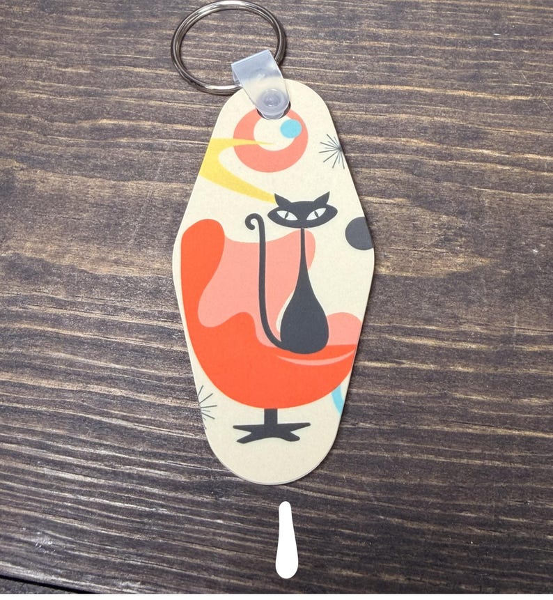 May include: A retro-style keychain featuring a black cat sitting in an orange chair. The keychain has a cream-colored background with a mid-century modern design, including a circular shape with yellow and blue accents. A silver key ring is attached.
