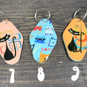 May include: Three retro-style cat keychains. The keychains have a diamond shape with a metal ring. One is peach with a black cat design, another is blue with cat faces, and the last is orange with a black cat design.