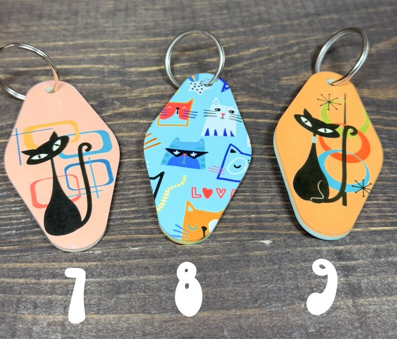 May include: Three retro-style cat keychains. The keychains have a diamond shape with a metal ring. One is peach with a black cat design, another is blue with cat faces, and the last is orange with a black cat design.